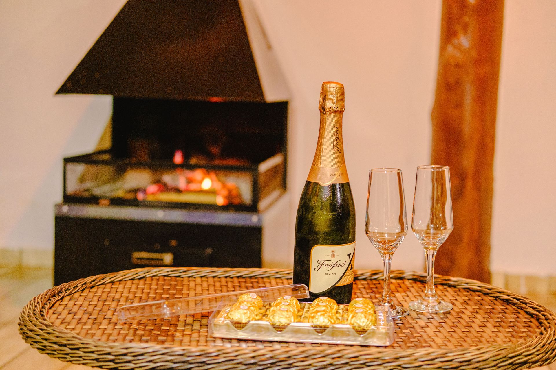 Garrafa de champanhe, taças e chocolates em uma bandeja; lareira ao fundo.