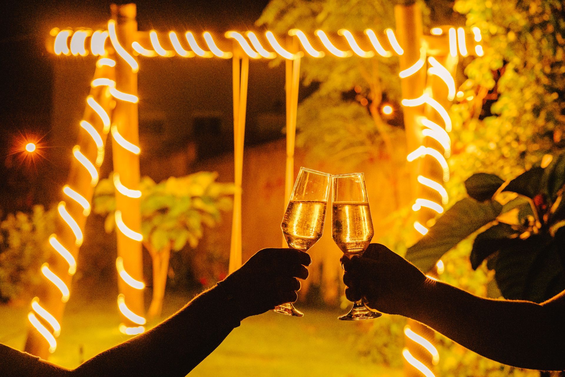 Mãos brindando com taças de champanhe em frente a um balanço de bambu iluminado por luzes de corda à noite.