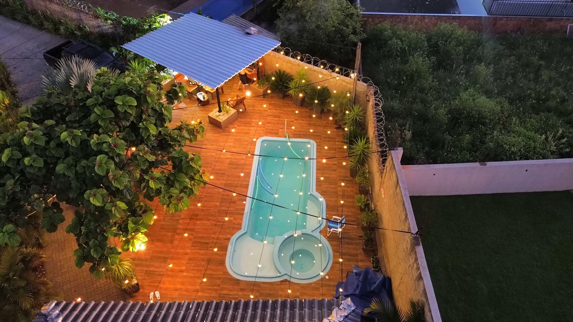 Área da piscina ao entardecer com luzes de corda, pátio de tijolos, assentos cobertos e vegetação.