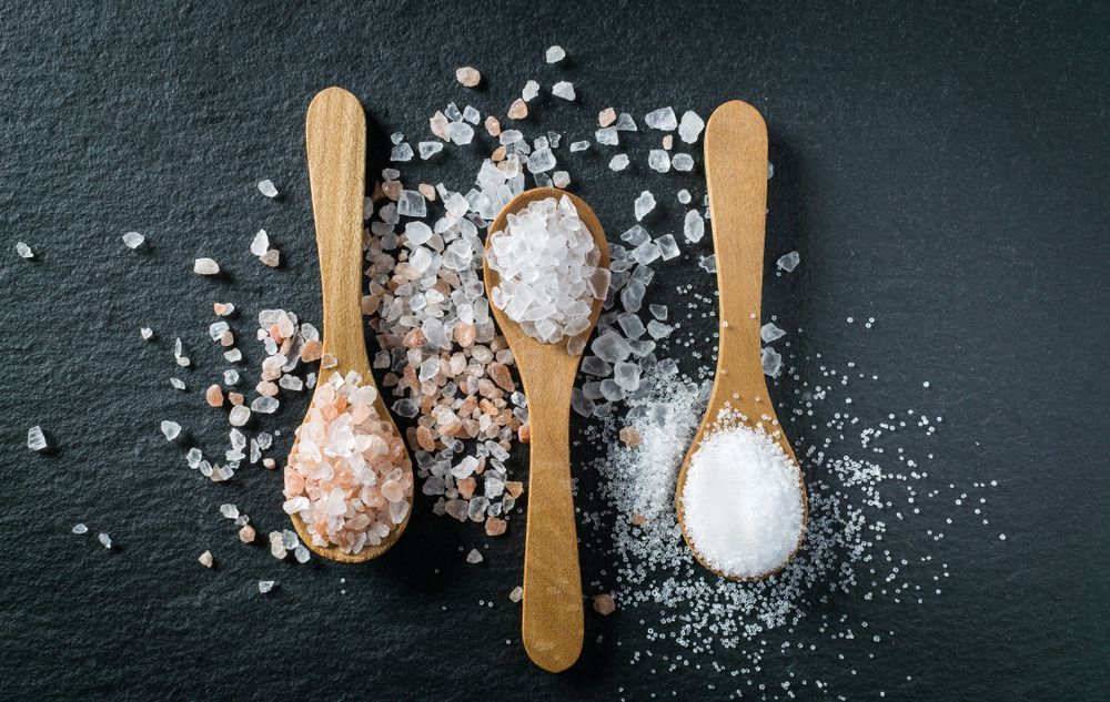 Three wooden spoons filled with different types of salt on a black surface.