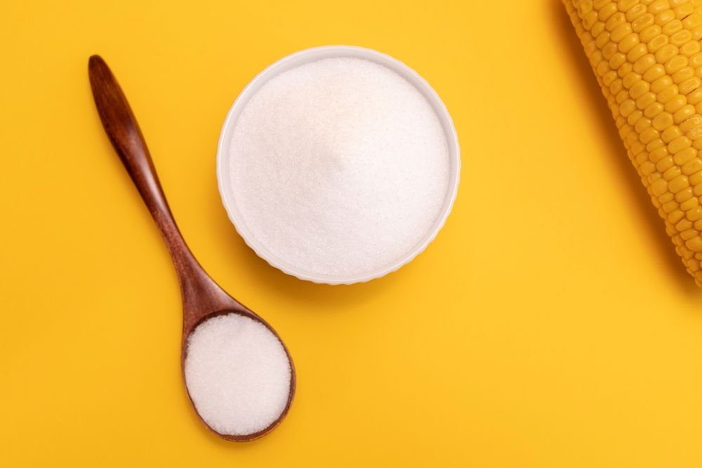 A bowl of sugar and a wooden spoon next to a corn on the cob on a yellow background.