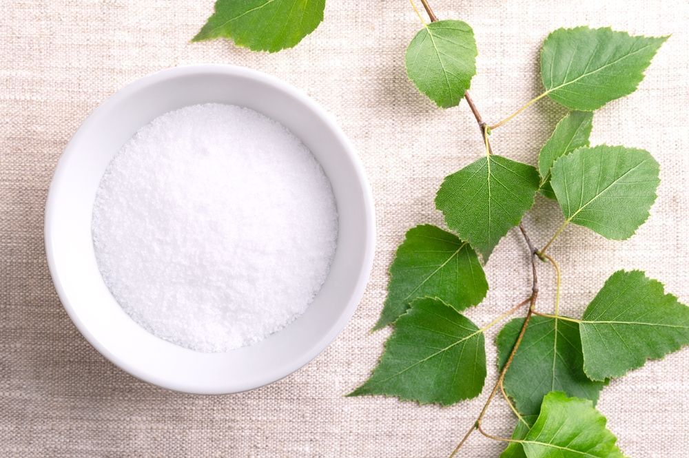 A bowl of xylitol next to a bunch of green leaves on a table.