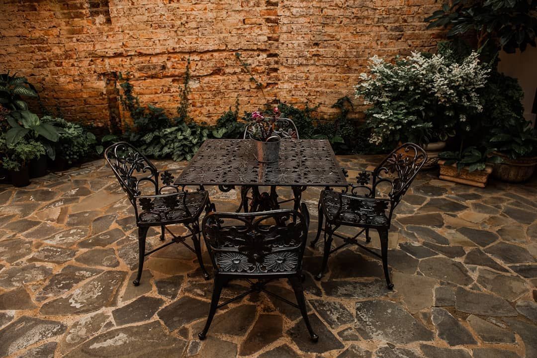 Mesa y sillas de hierro forjado en un patio de piedra, con una pared de ladrillos y plantas al fondo.