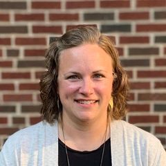 Woman with light brown hair smiles at the camera in front of a brick wall.