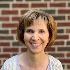 Woman with short, light brown hair smiles in front of a brick wall, wearing a light-colored sweater.