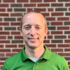 Man with light skin smiling, wearing a green polo shirt, in front of a brick wall.
