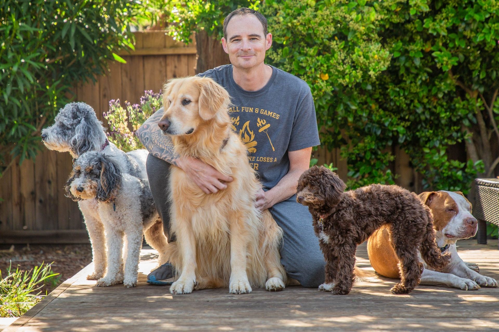 Man kneels with five dogs of various breeds on a wooden deck outdoors; sunny.
