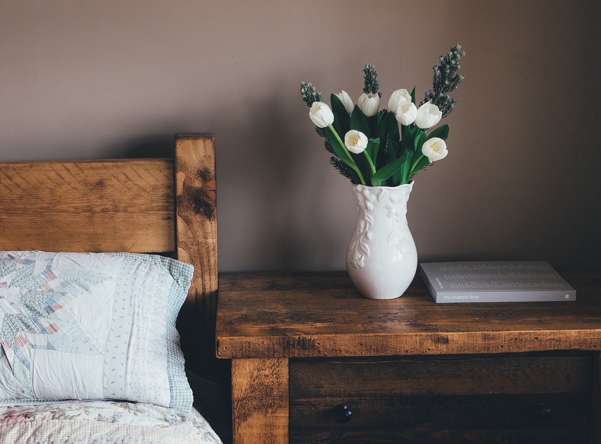 A vase of flowers is sitting on a wooden nightstand next to a bed.