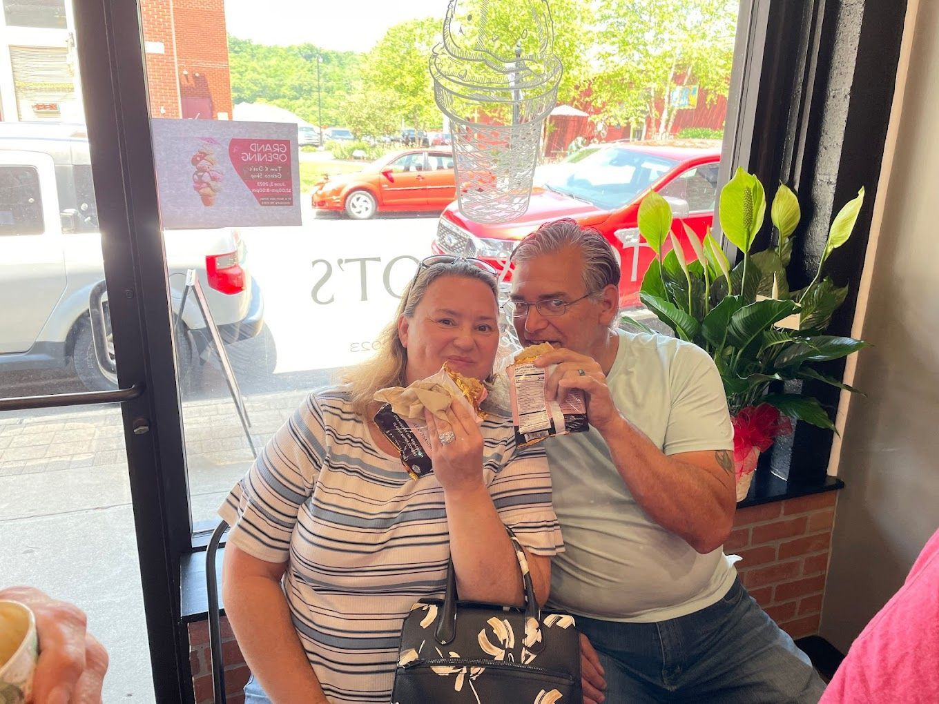 A man and a woman are sitting in front of a window eating ice cream sandwiches.
