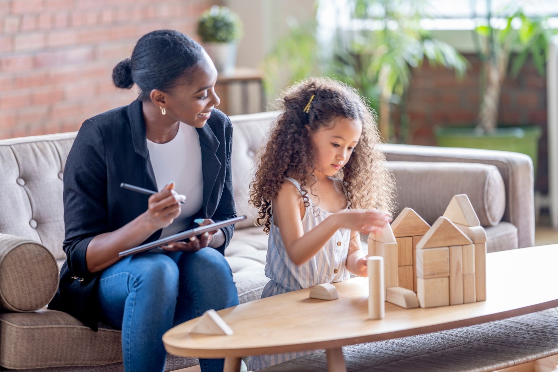 A woman is sitting on a couch talking to a little girl who is playing with wooden blocks.