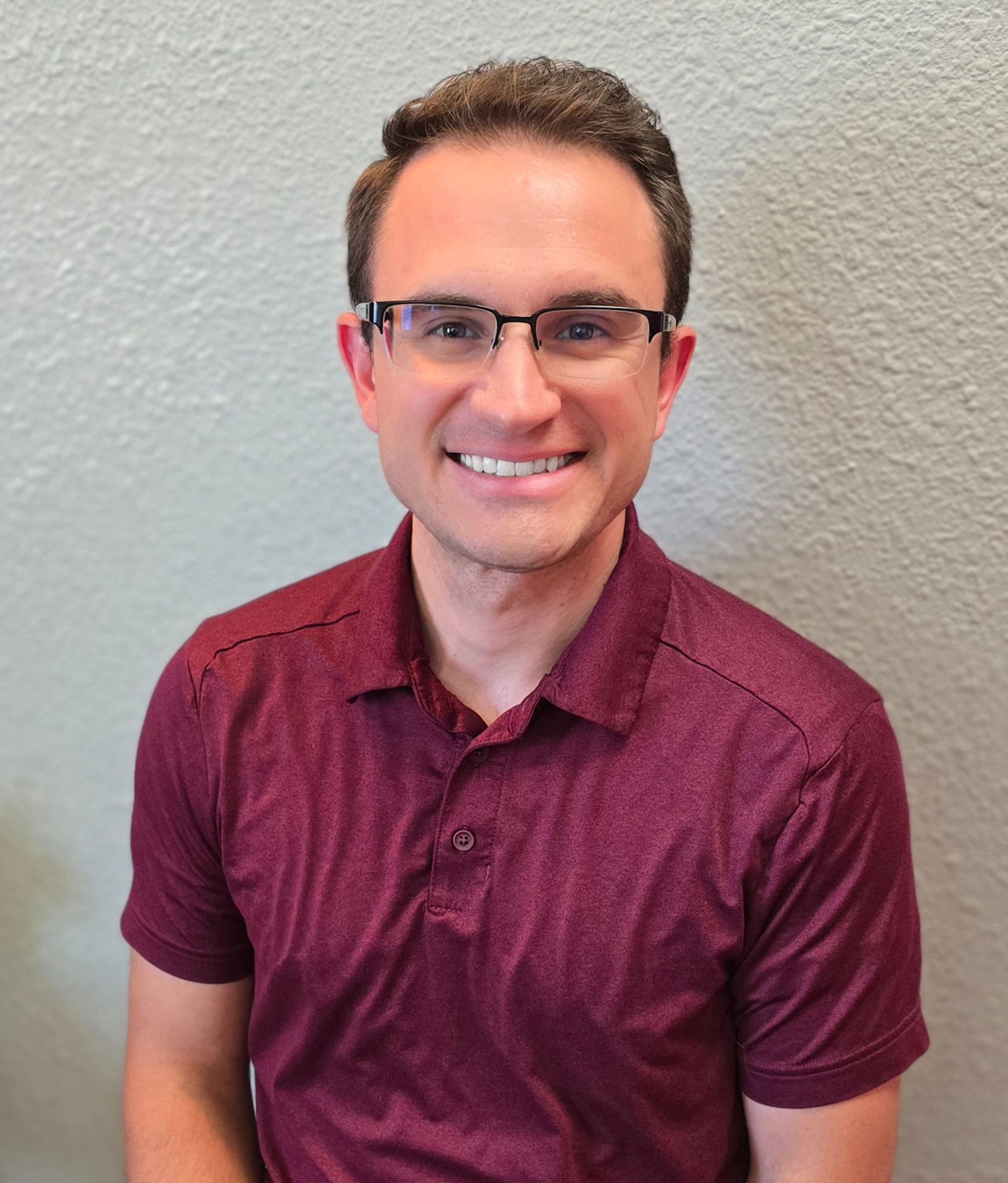A man wearing glasses and a maroon shirt is smiling for the camera.