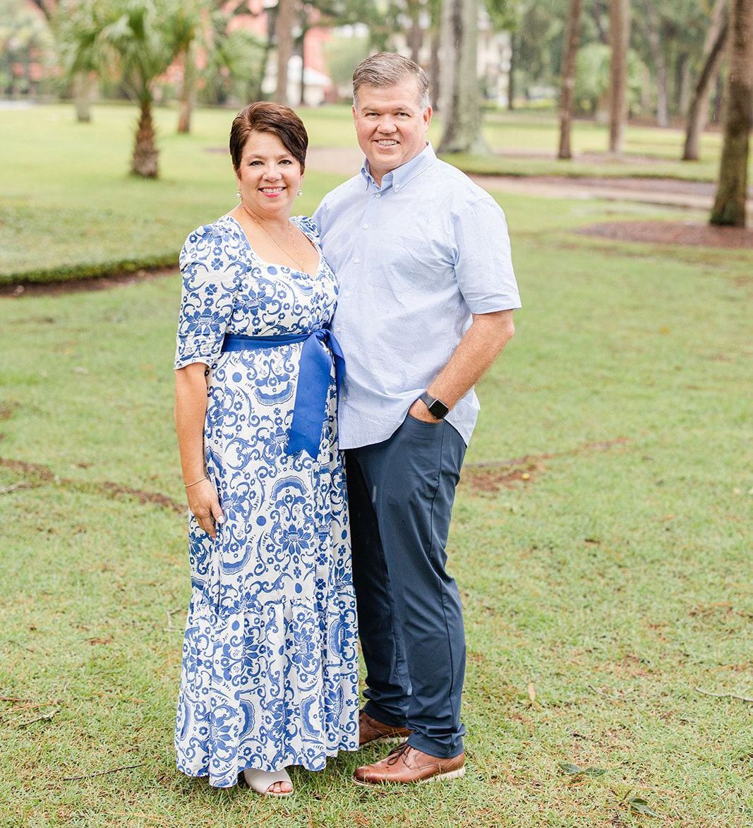 Couple poses outdoors. Woman in blue and white dress, man in light blue shirt and navy pants.