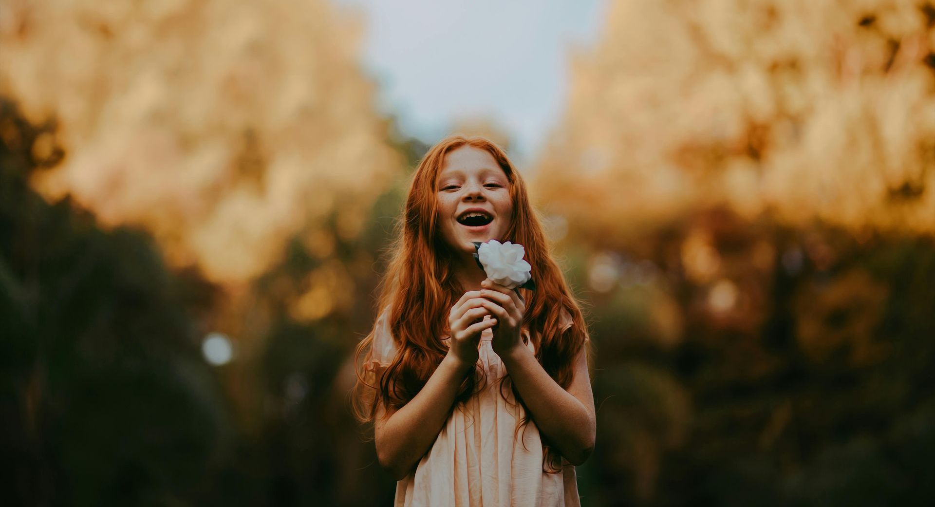 A young girl with red hair is holding a flower in her hands and smiling.
