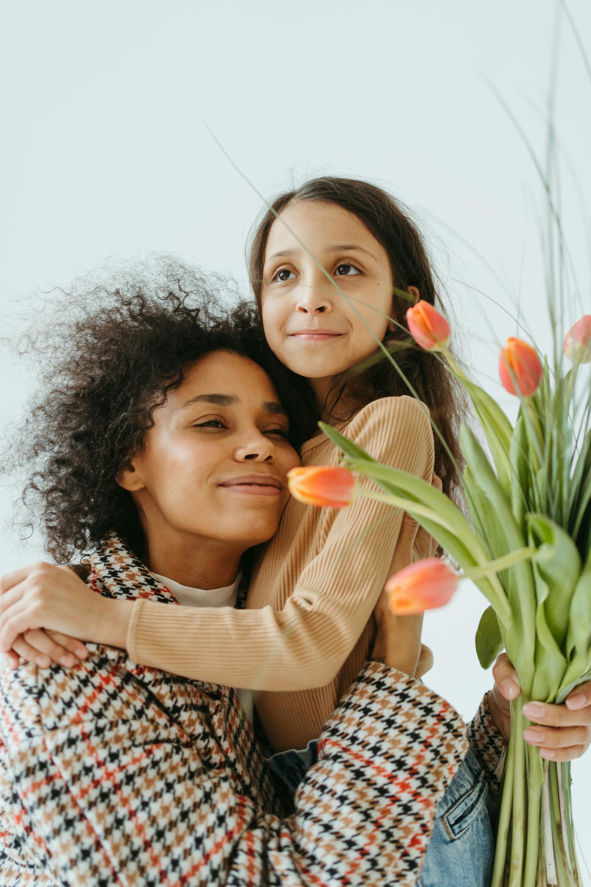 Mother and daughter embrace, holding tulips. The daughter smiles, and the mother is joyful. White background.