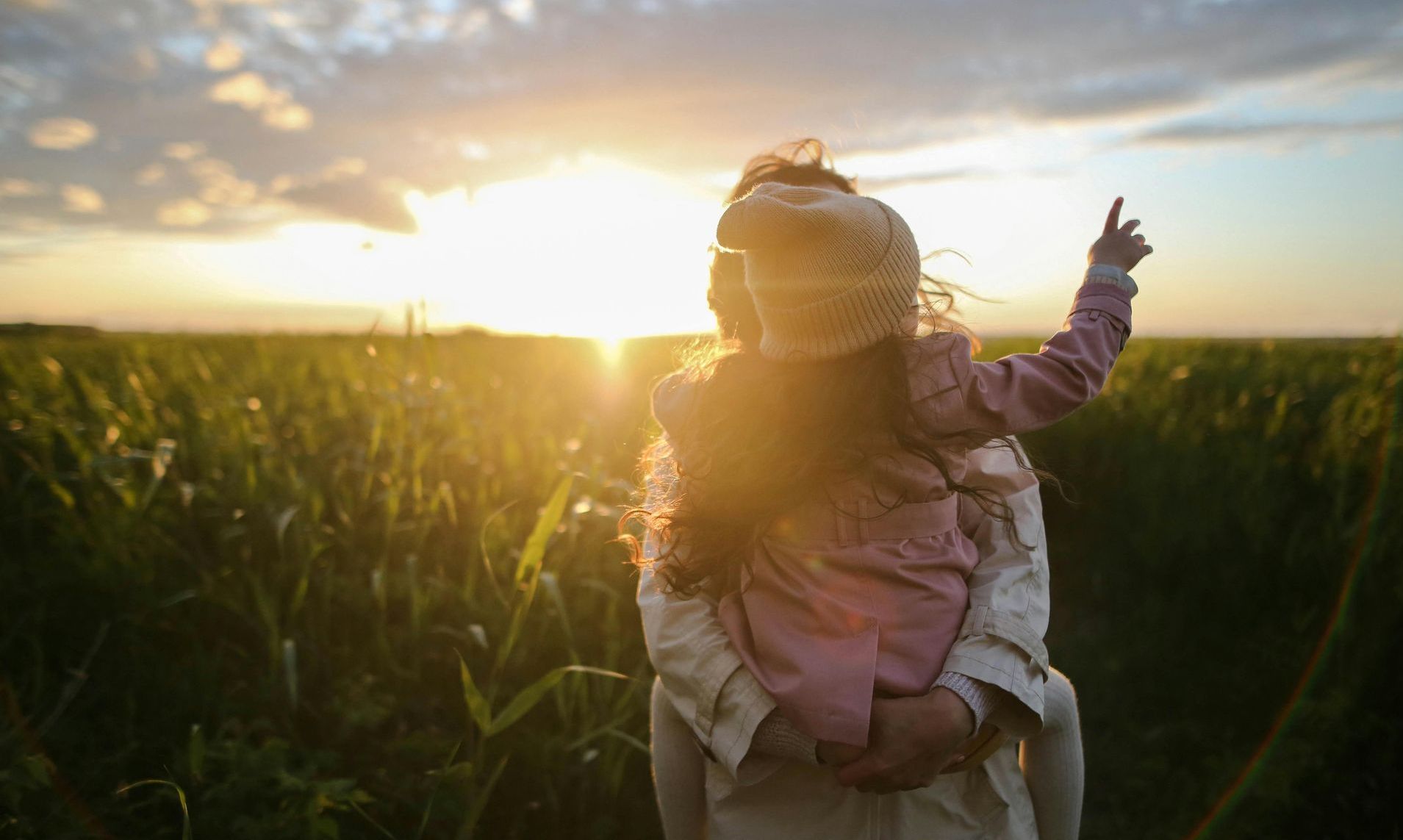 A woman is holding a little girl on her shoulders in a field at sunset.