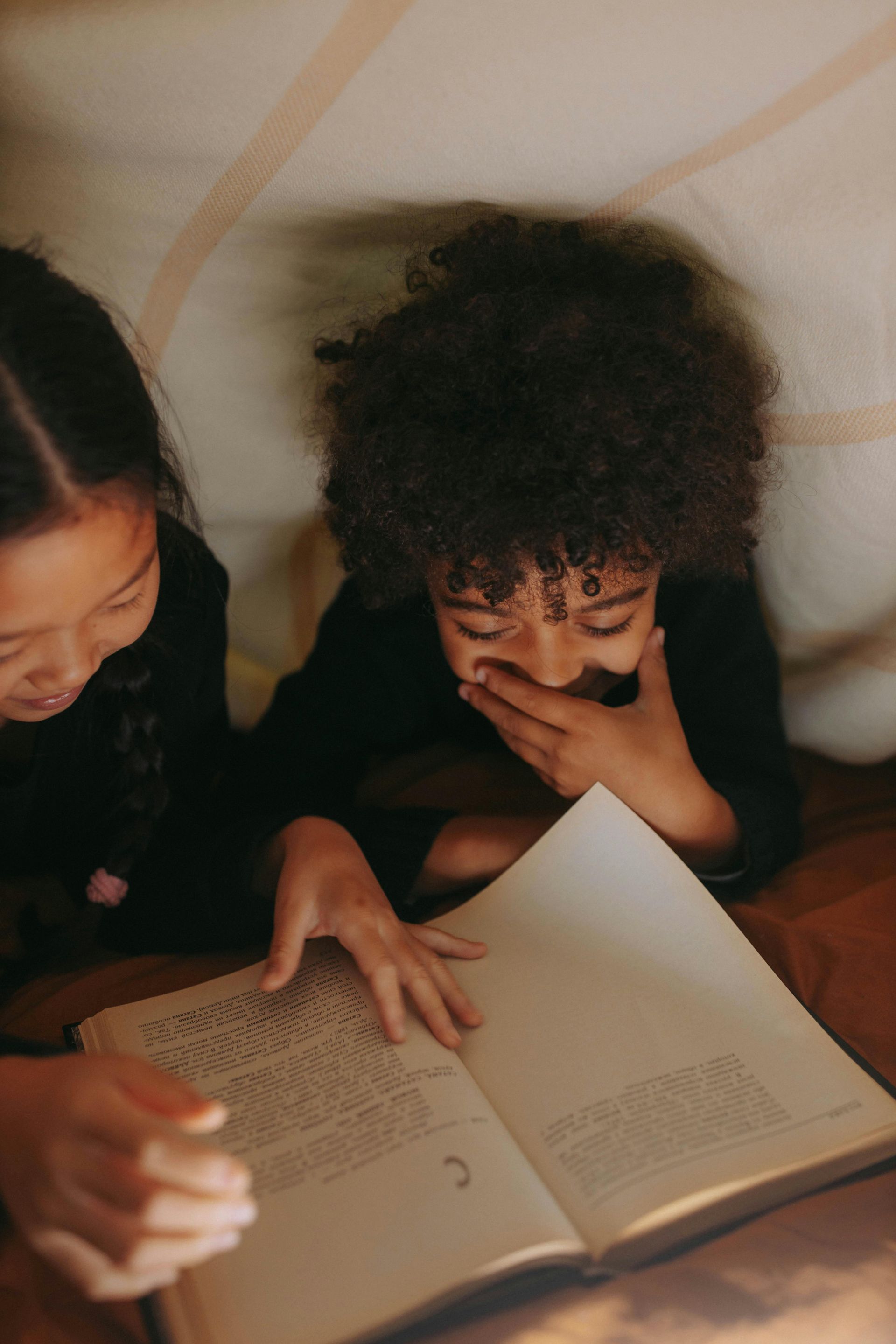 A boy and a girl are reading a book under a blanket.