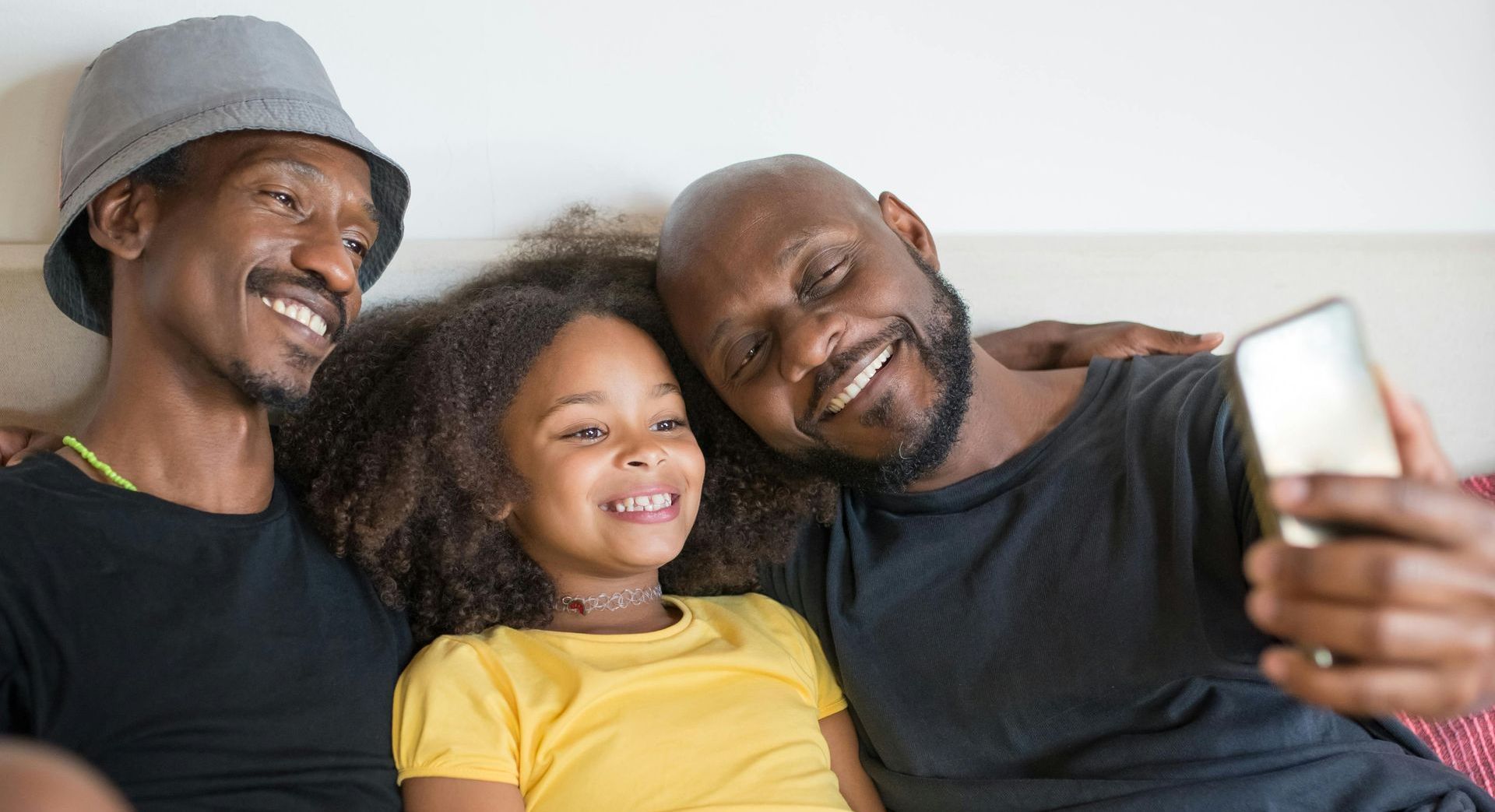 Two men and a little girl are sitting on a couch taking a selfie with a cell phone.