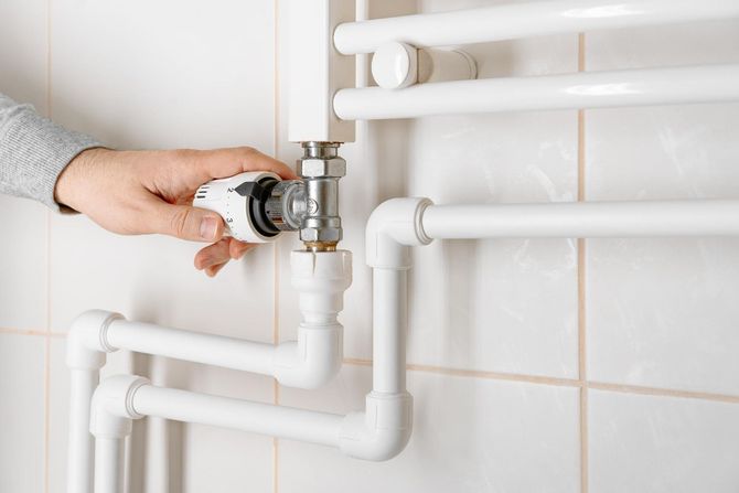 Person's hand adjusting a white radiator valve on a white towel rack in a tiled bathroom.