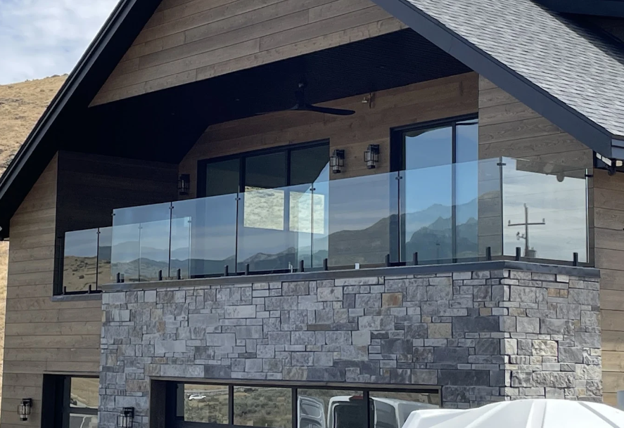 Balcony with glass railings on a house with stone and wood siding, overlooking mountains.