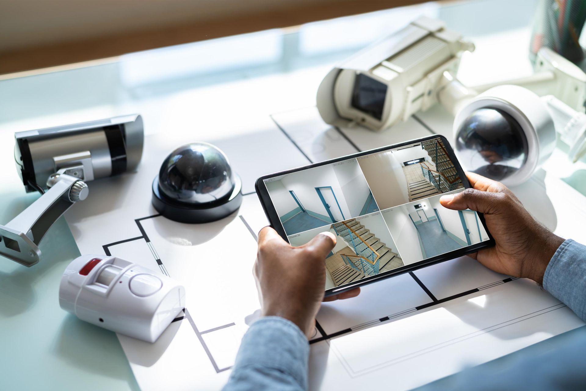 Hands holding tablet displaying security camera views, surrounded by security cameras and blueprints.