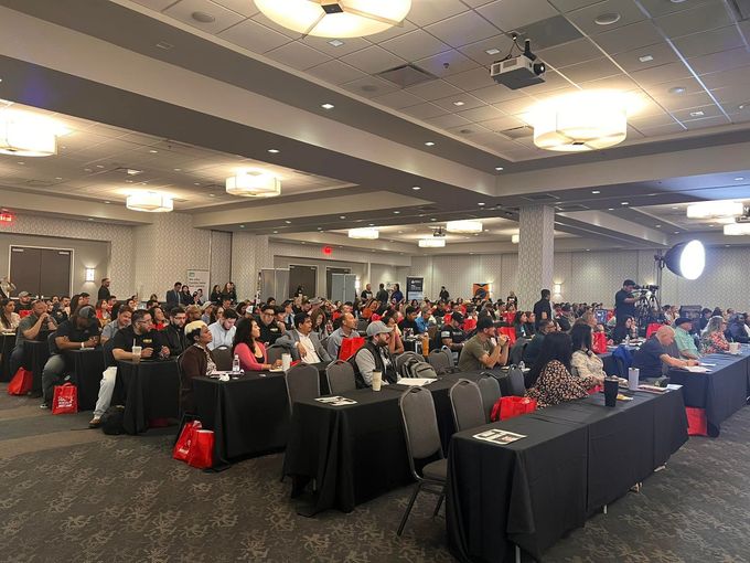 Large conference room filled with people seated at tables, listening to a presentation.