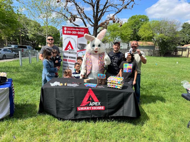 People and a bunny mascot at a table for Apex Smart Home. Displayed goods and a sign are visible. Outdoor setting.