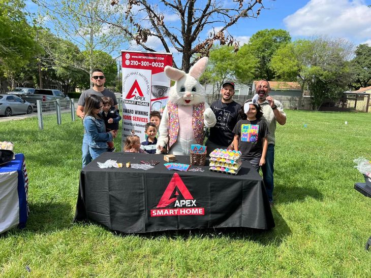 People and a bunny mascot at a table for Apex Smart Home. Displayed goods and a sign are visible. Outdoor setting.