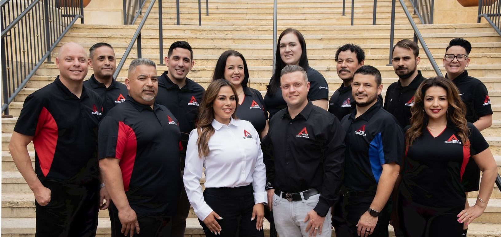 A group of people wearing black shirts with a logo, standing on stairs. Some have red accents, a woman wears white.