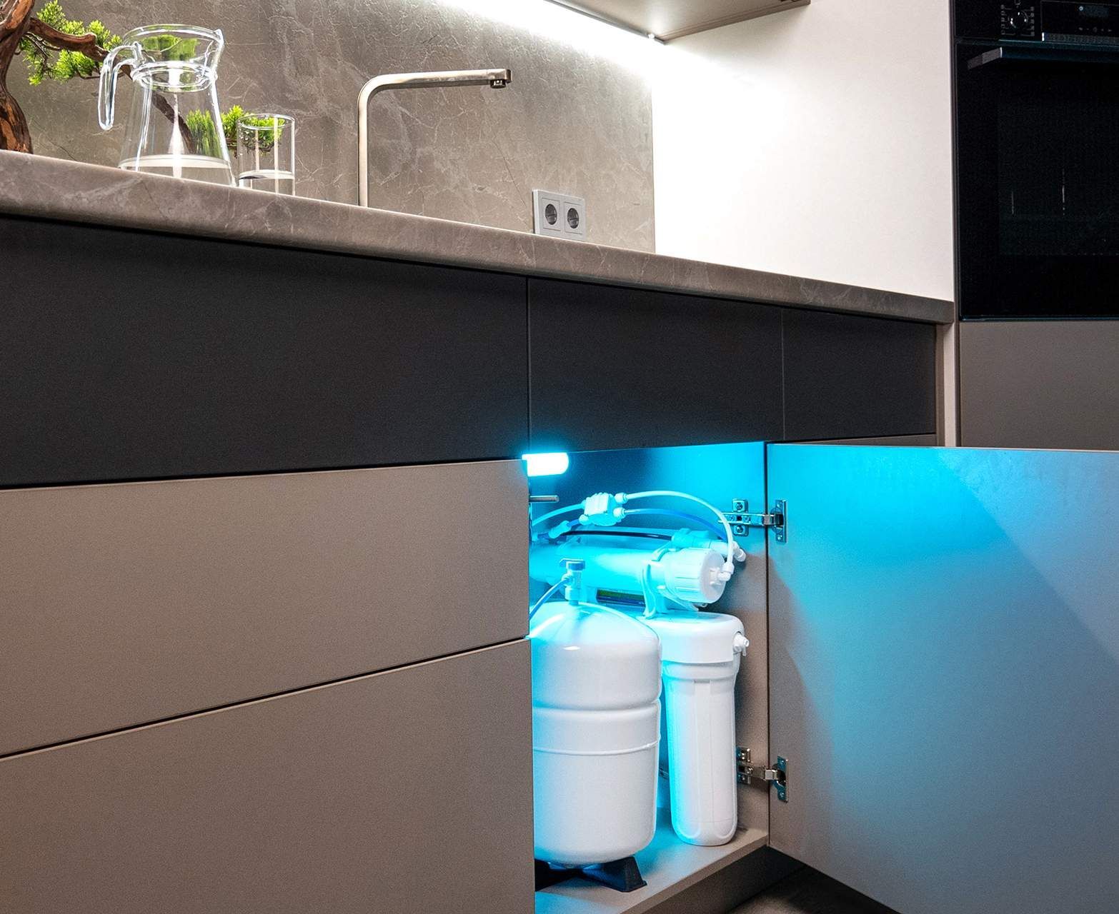 Kitchen cabinet with an open door, revealing a water filtration system illuminated by blue light.