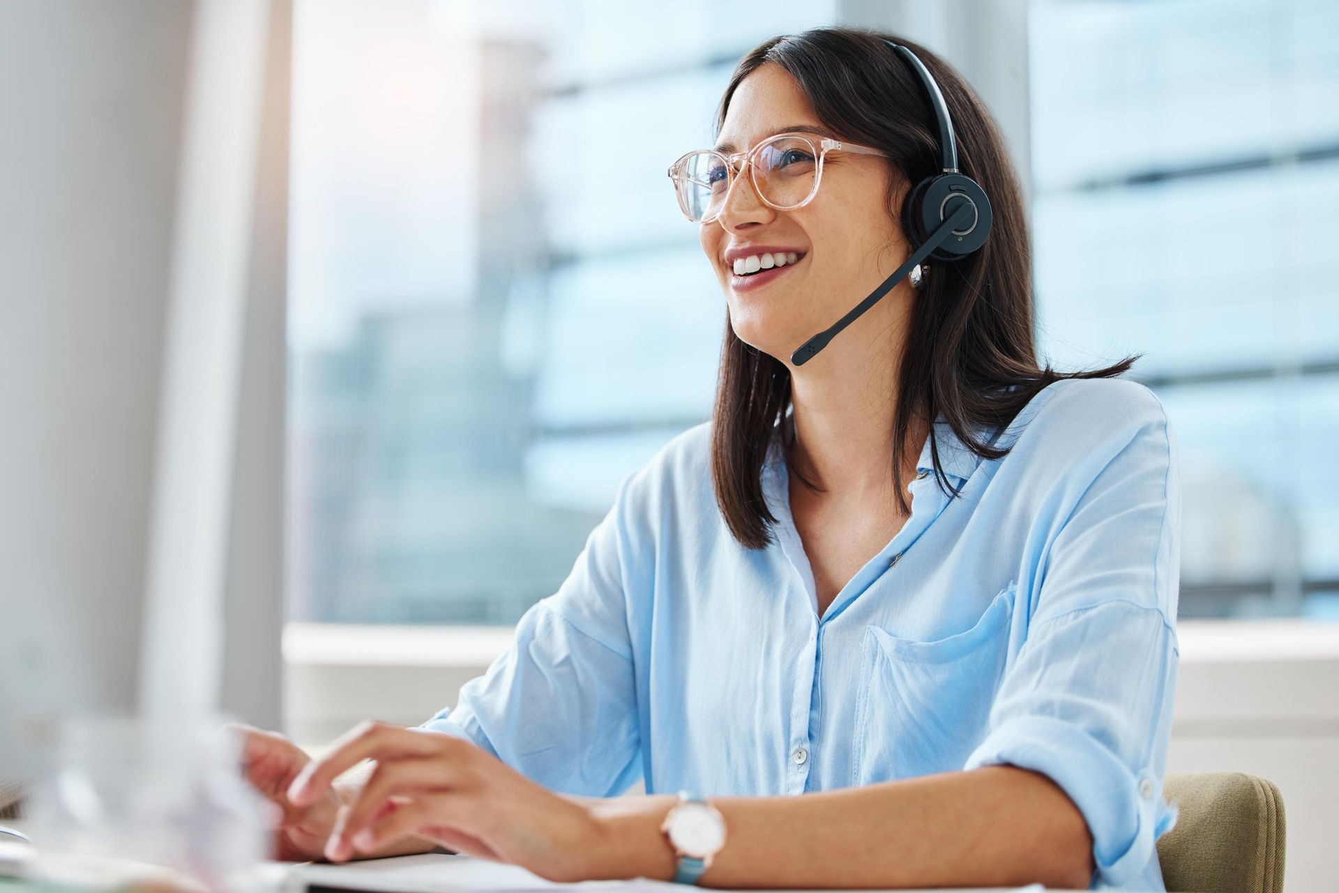 Woman wearing headset smiling at a computer, sunny office setting.