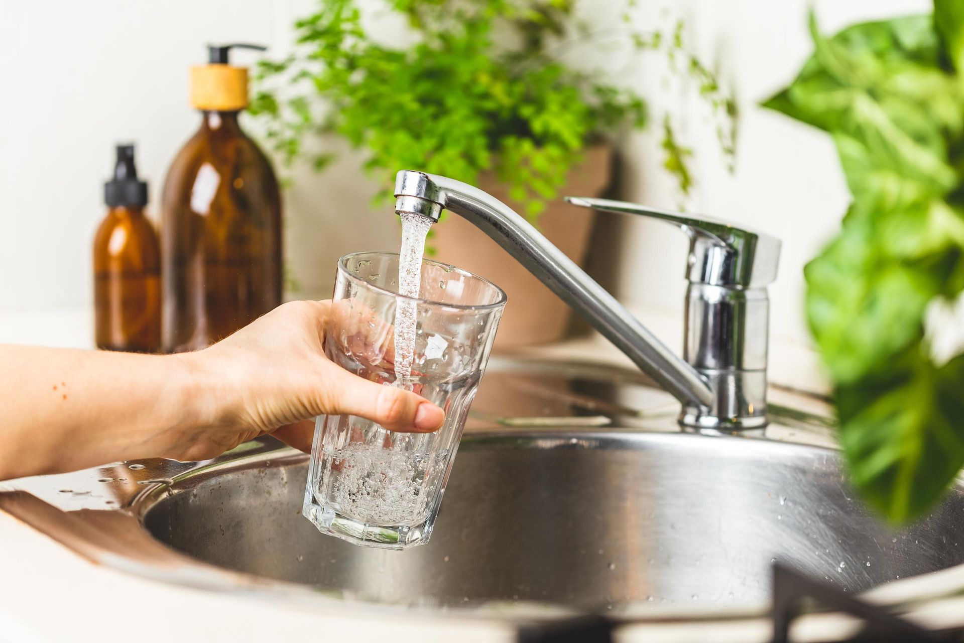 Hand filling a glass with water from a kitchen faucet, plants and soap bottles in background.