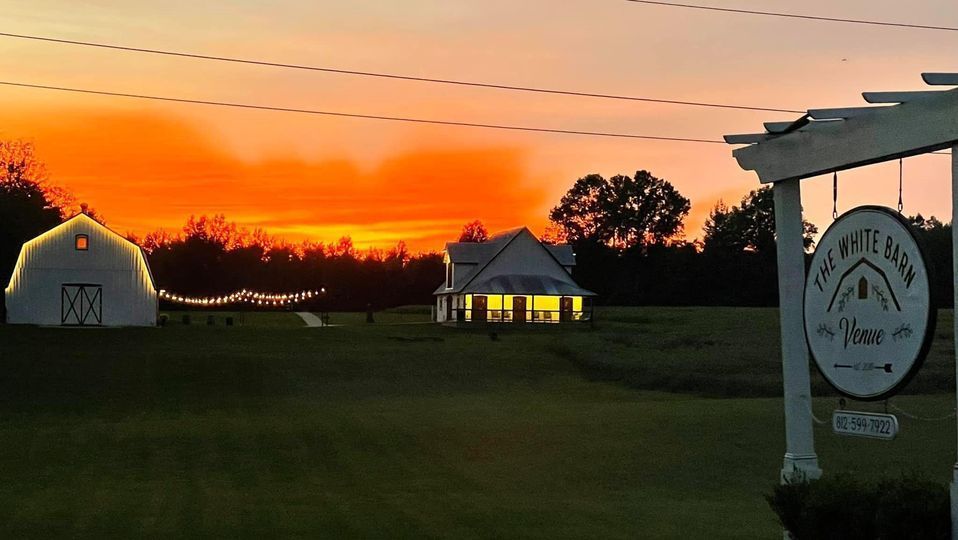 Sunset over a rural property with a white barn, house, and sign for 