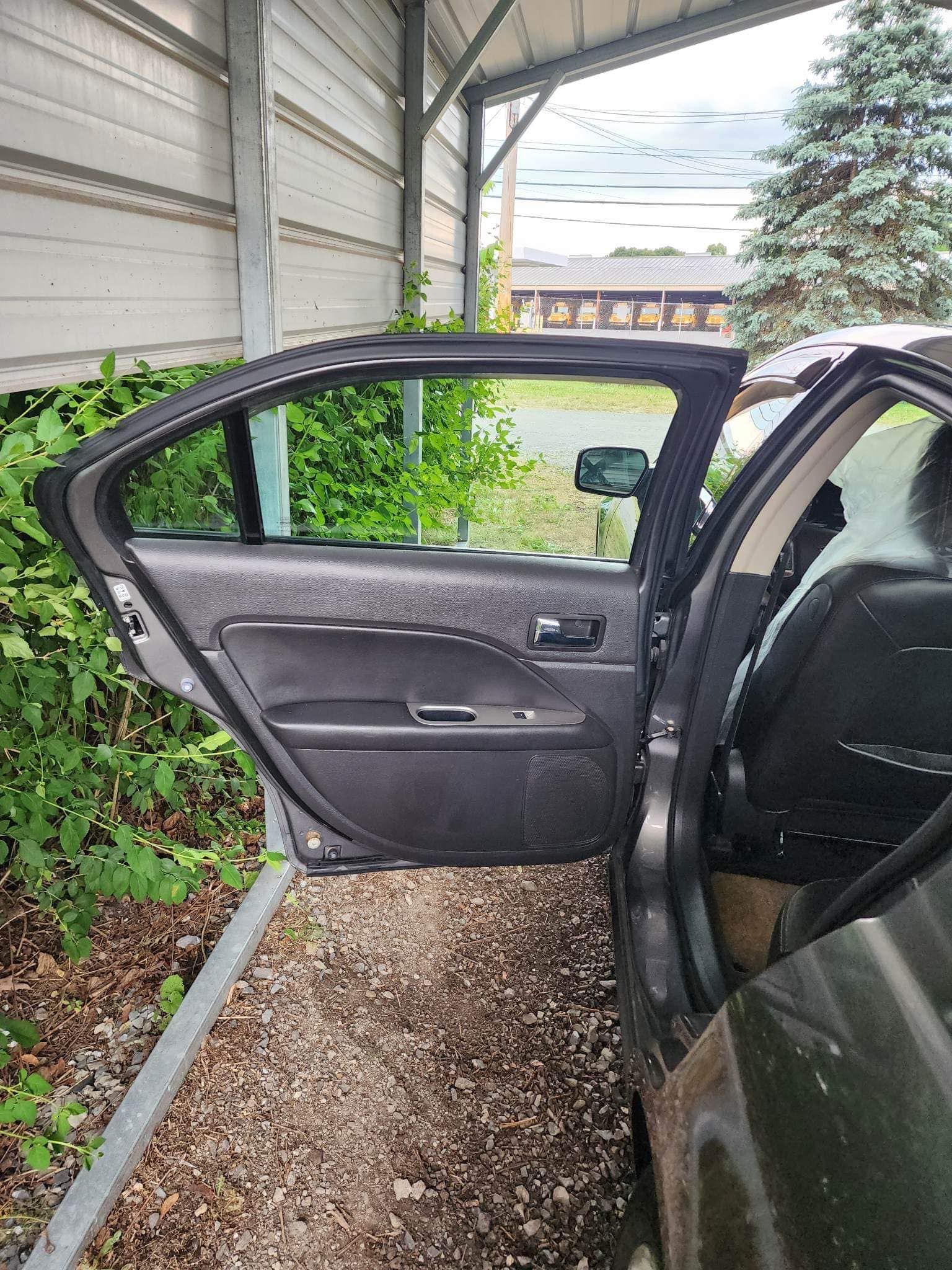 Open car door, showing interior and surrounding outdoor area with vegetation and a shelter.
