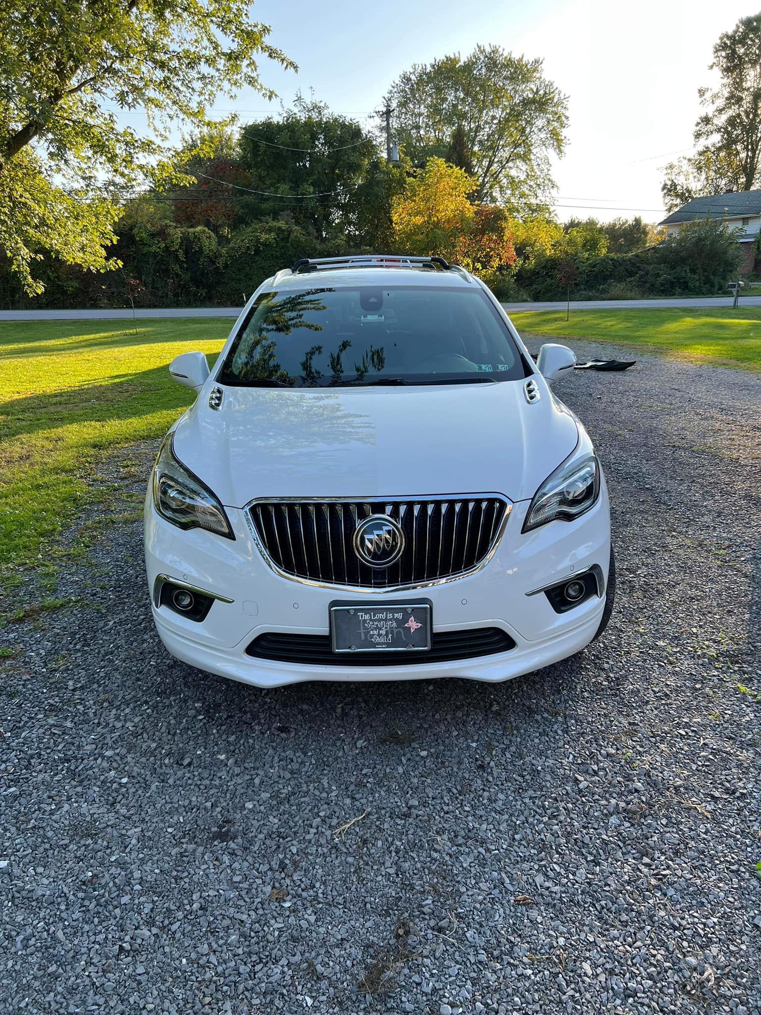 White Buick SUV parked on gravel driveway, with green grass and trees in the background.