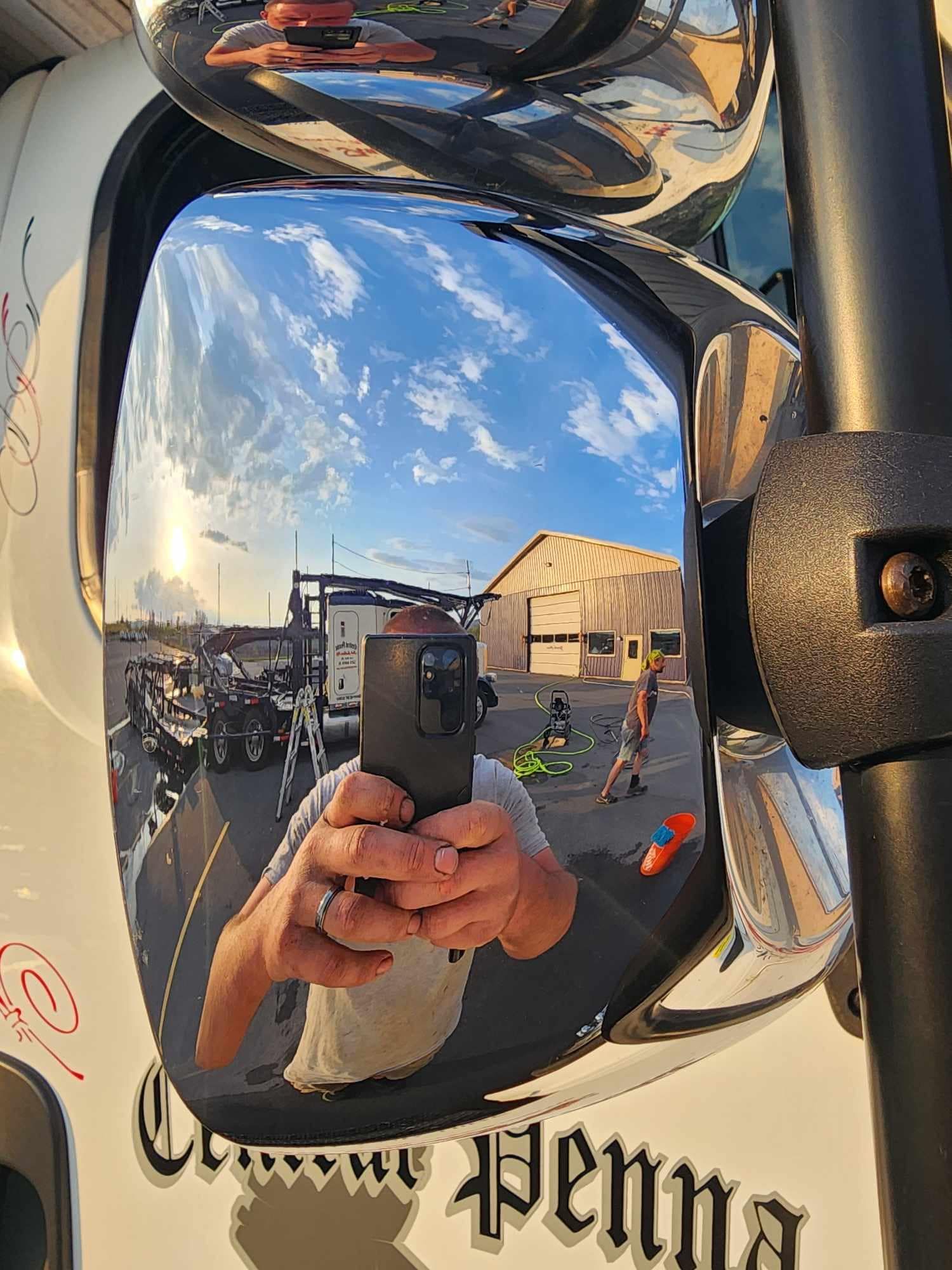 Reflection in a truck mirror showing a person taking a photo of a sunny scene with other vehicles.