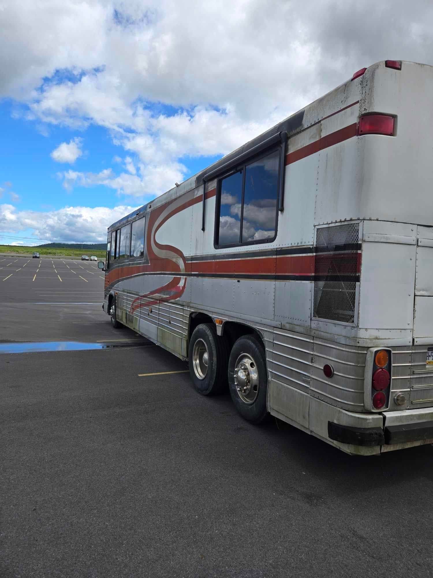 A silver and white bus with red stripes on a gray asphalt surface, against a cloudy blue sky.