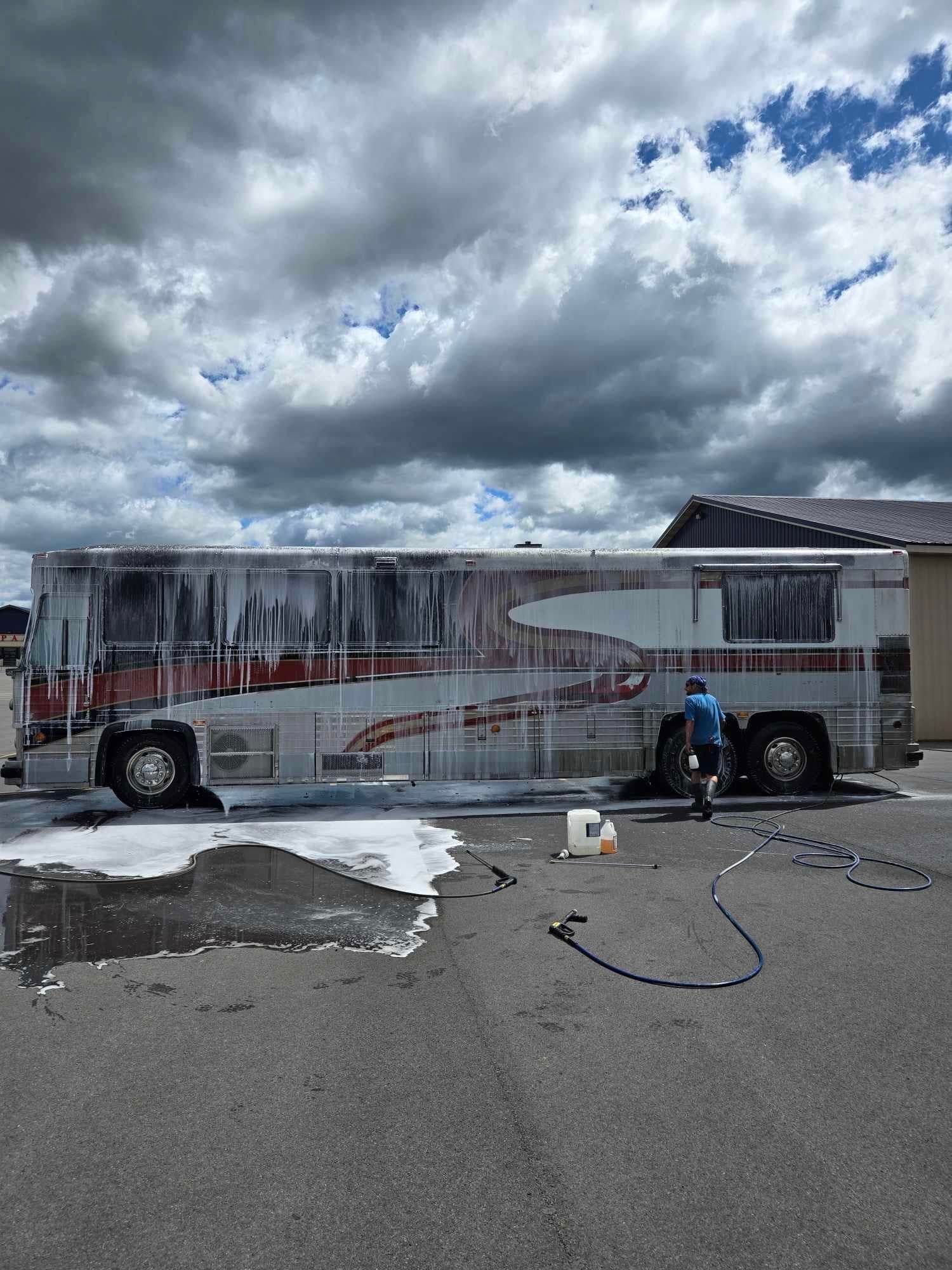 Person washing a large red and white bus with white foam, outside on a cloudy day.