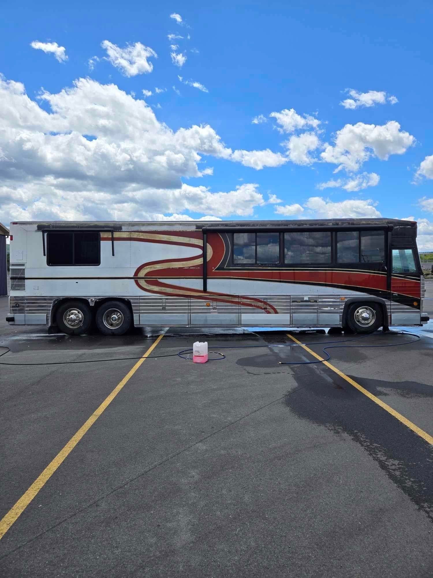 A large, vintage bus with red and gold accents parked on asphalt, bathed in sunlight under a blue sky.