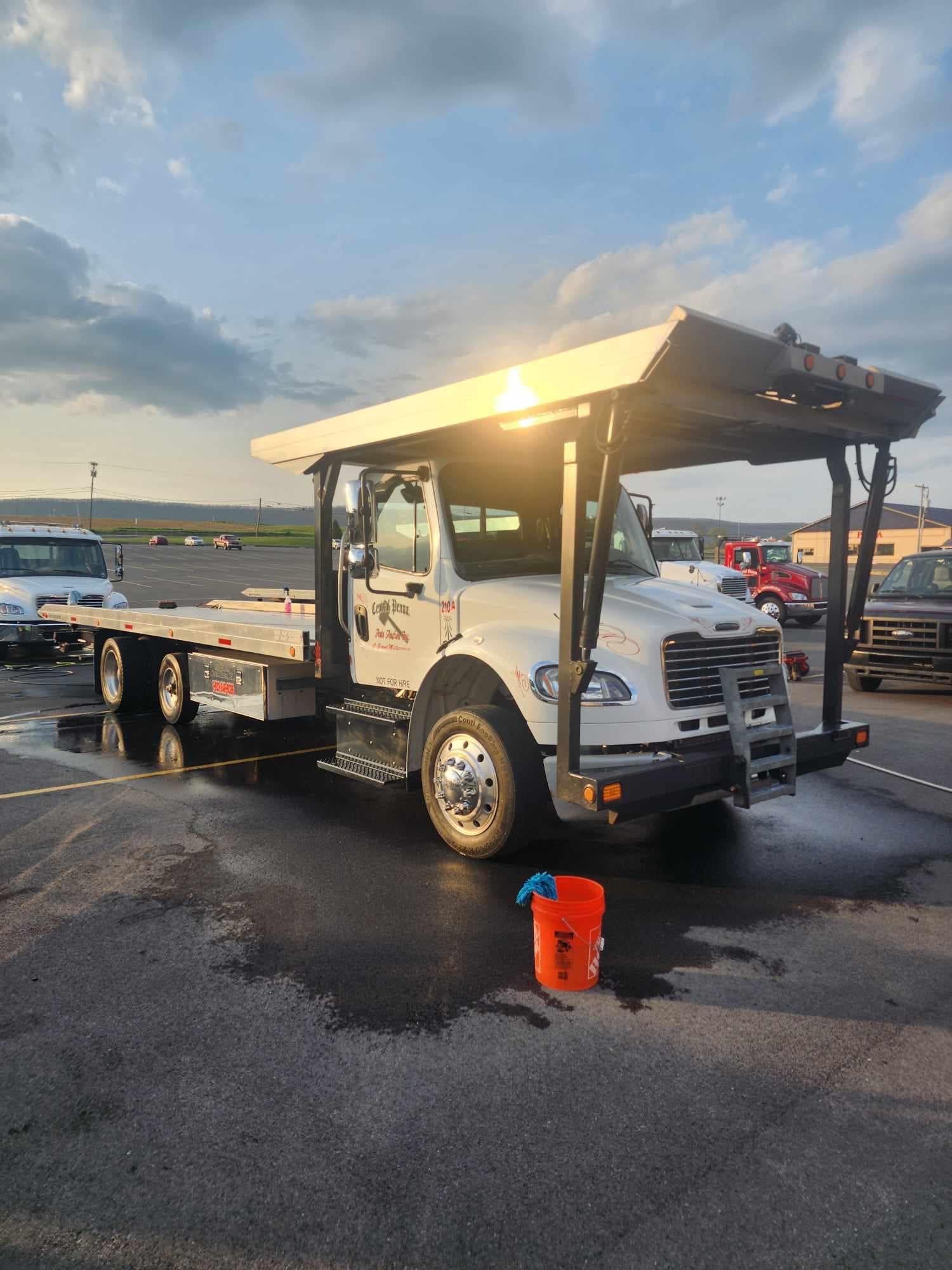 White tow truck with an open flatbed, parked on wet asphalt, with a bucket in front.