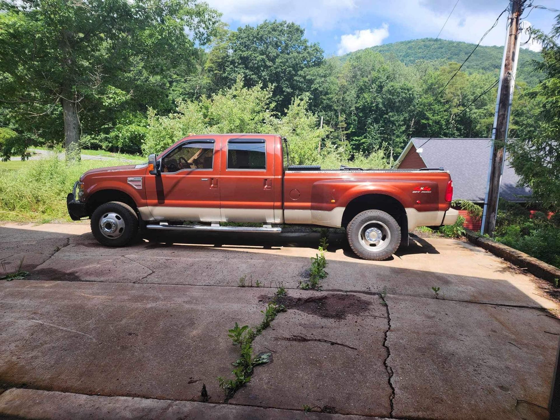 Brown and tan pickup truck parked on cracked concrete in front of a house and trees.