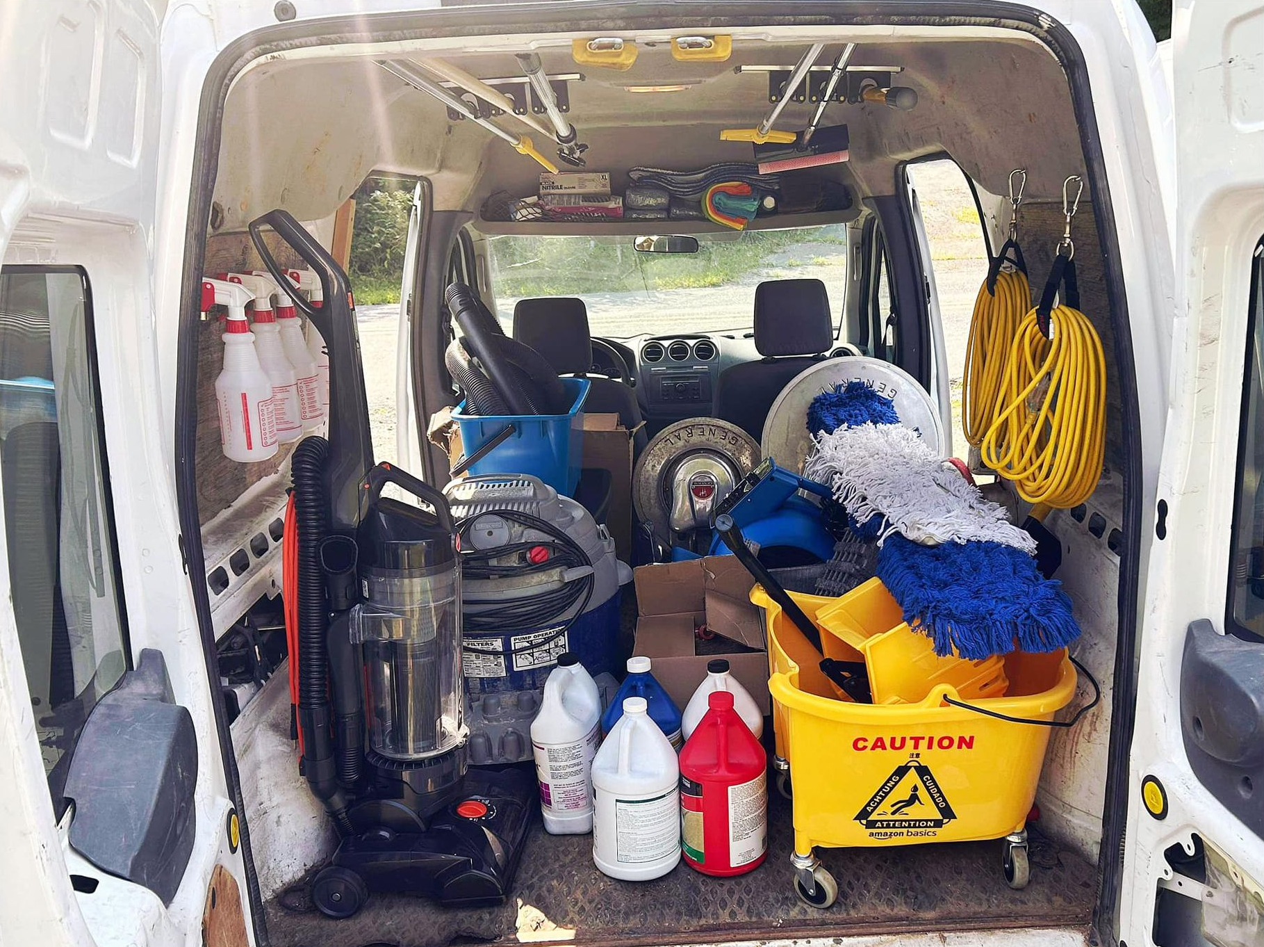 Open white van interior filled with cleaning supplies: vacuum, buckets, mops, and cleaning liquids.