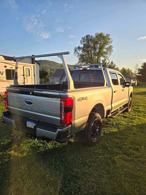 Silver Ford pickup truck with ladder rack parked on grass. A camper is in the background.
