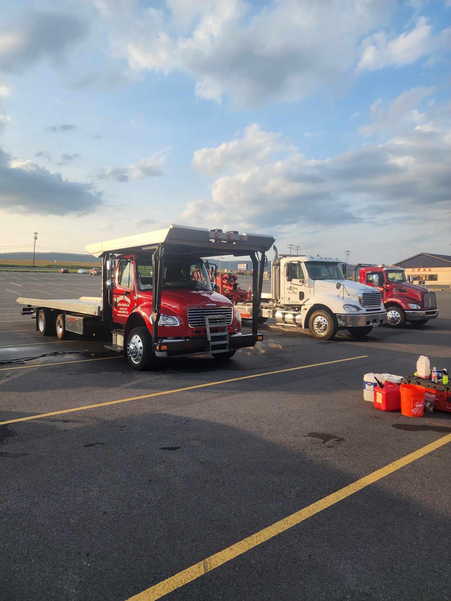 Red, white, and red tow trucks parked on asphalt under a cloudy sky. Buckets sit nearby.