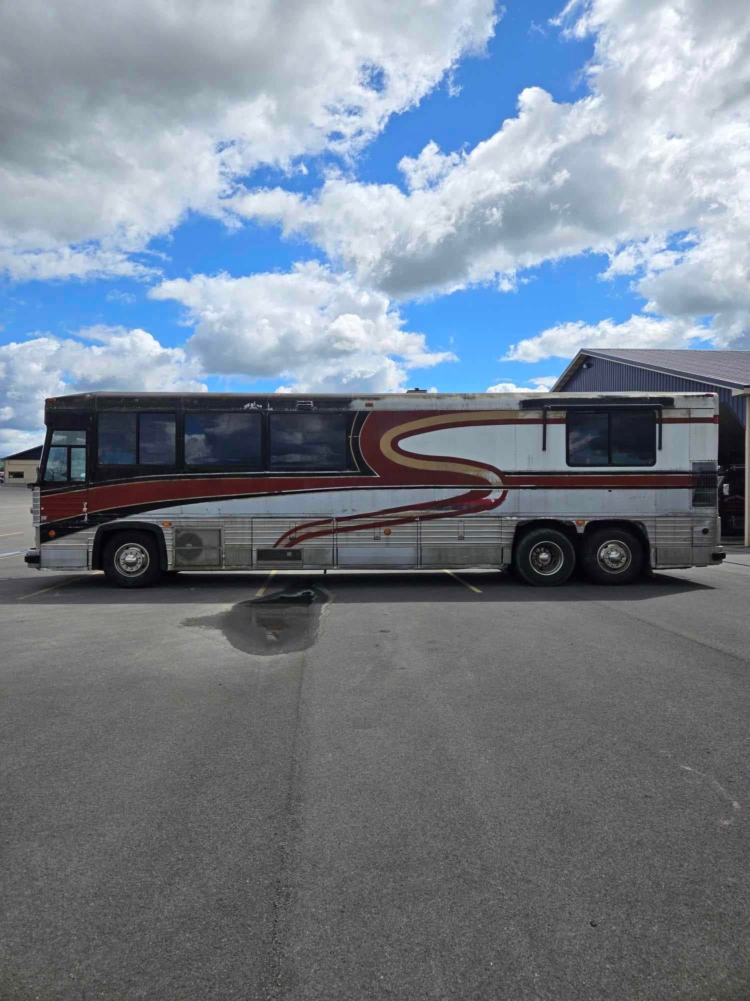 A large, silver and red bus with a stylized brown stripe is parked on asphalt under a cloudy sky.