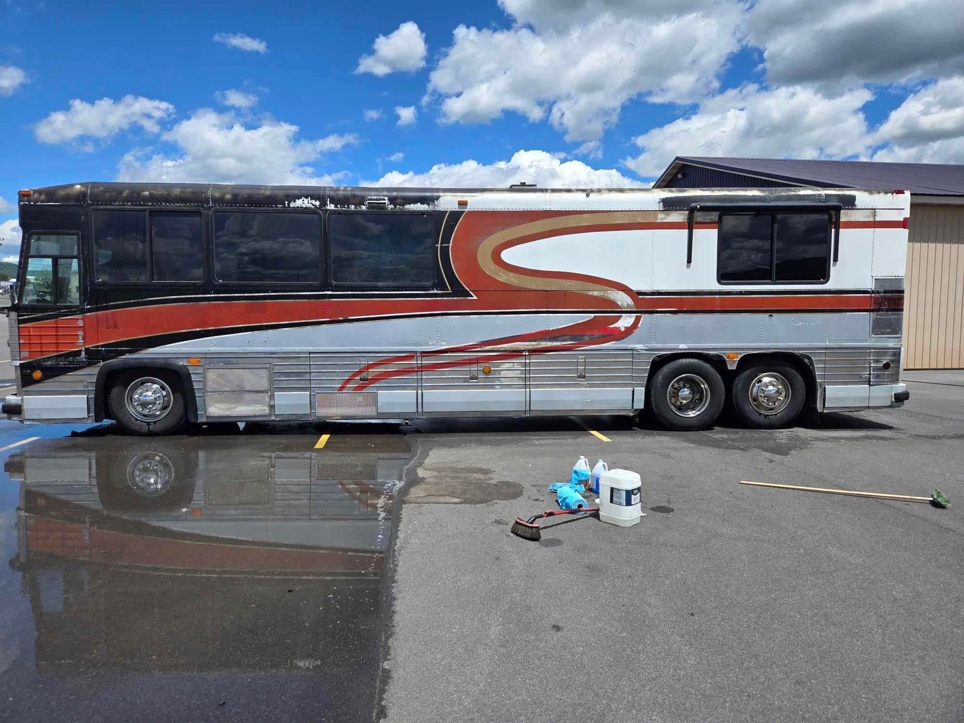 Side view of a weathered, white, orange, and silver bus parked on wet pavement under a blue sky.