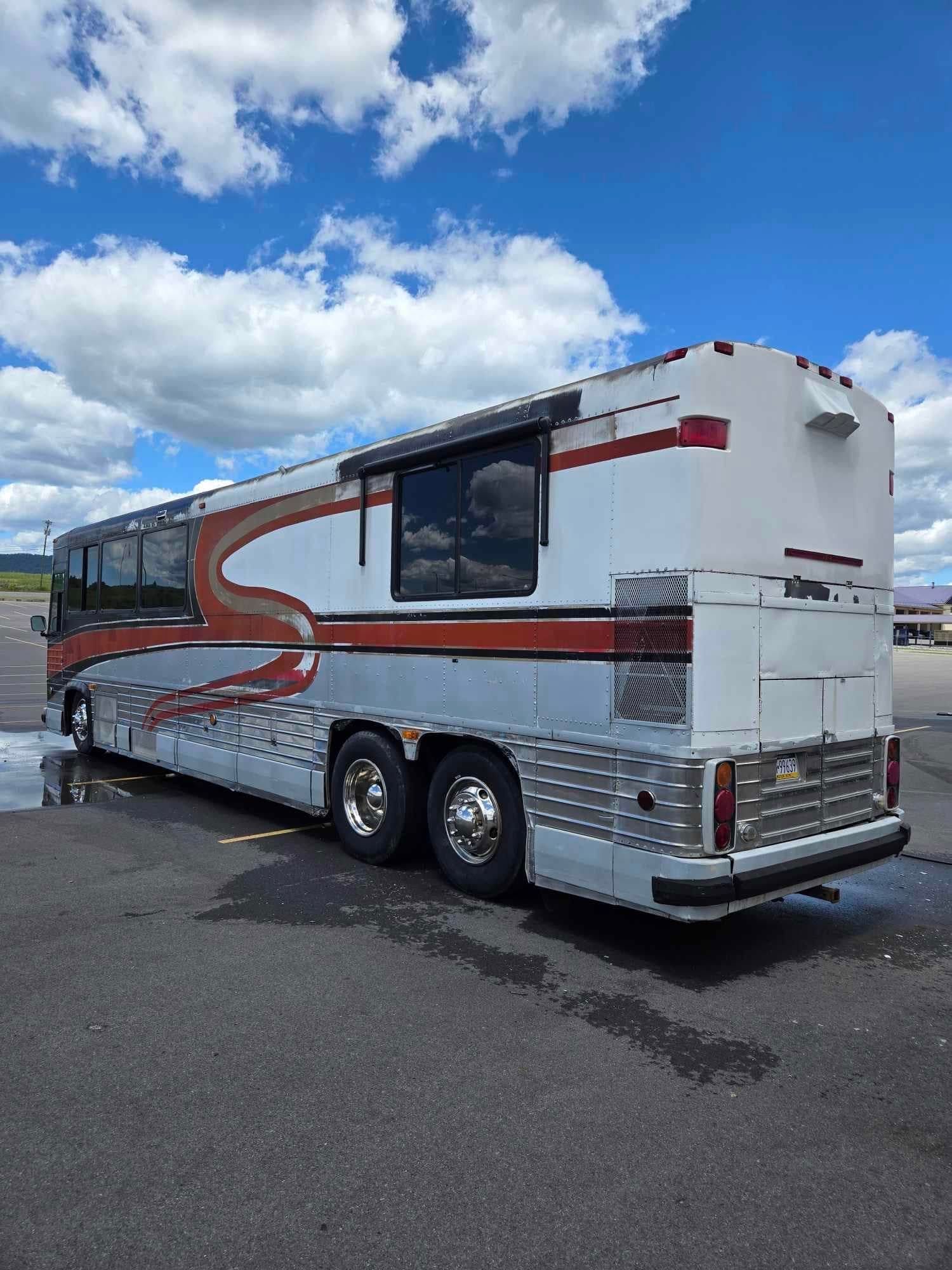 RV-style bus, white with silver and red accents, parked on asphalt under a partly cloudy blue sky.