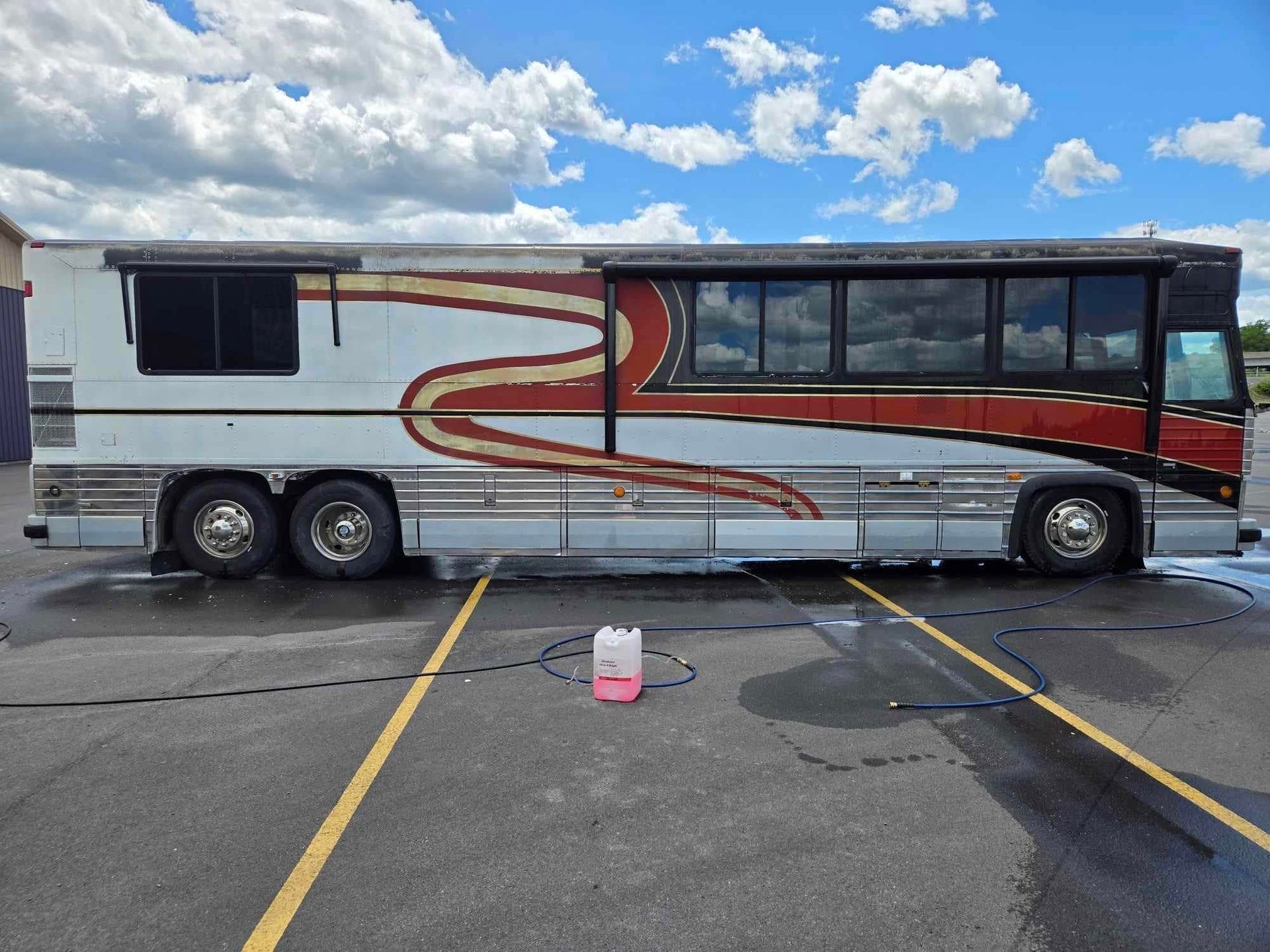 A large, weathered bus being washed outdoors, with a white and red color scheme, parked on a gray asphalt surface.