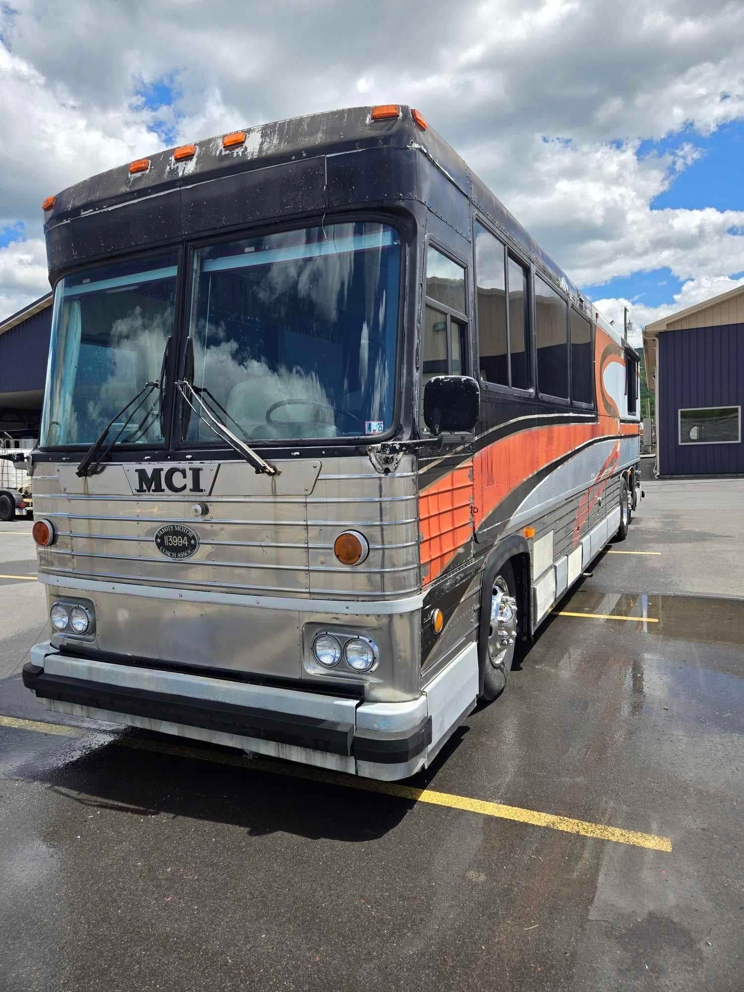 A large, silver MCI bus with orange and black accents parked on wet asphalt under a cloudy sky.