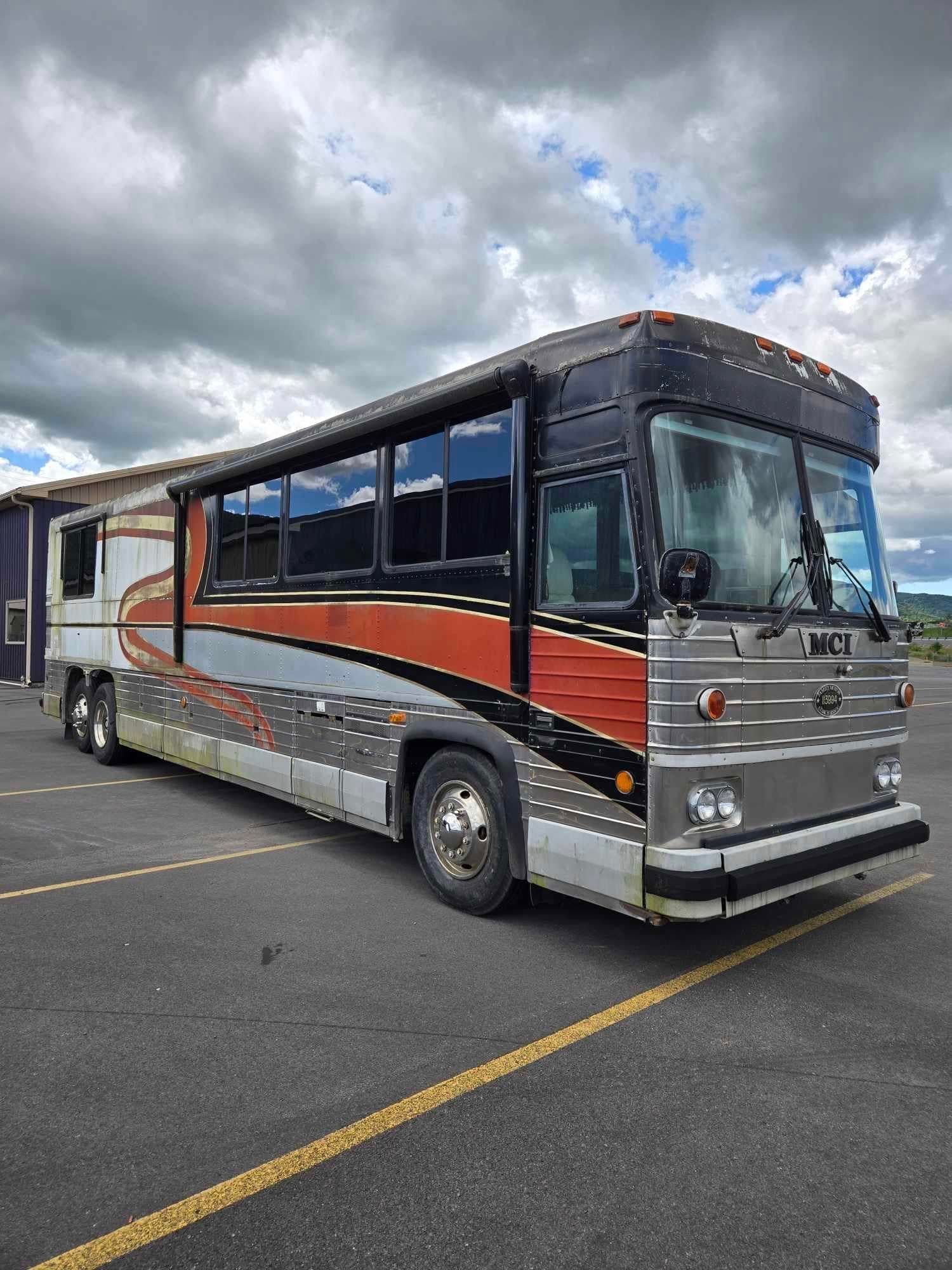 Gray, orange, and black bus on asphalt under a cloudy sky.