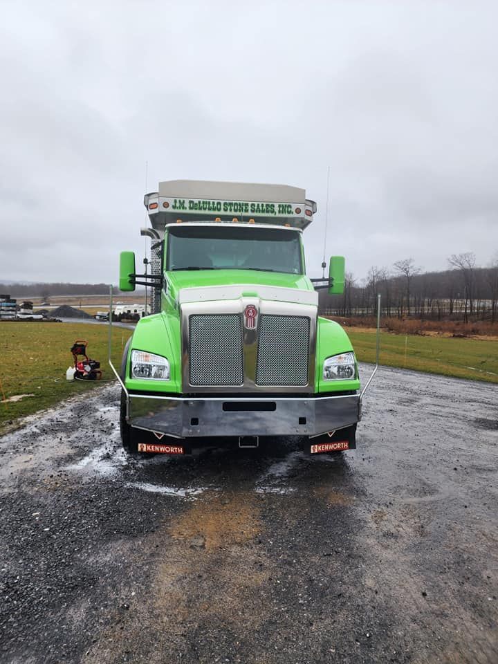 Green Kenworth dump truck on a wet road, chrome grill, under cloudy sky.